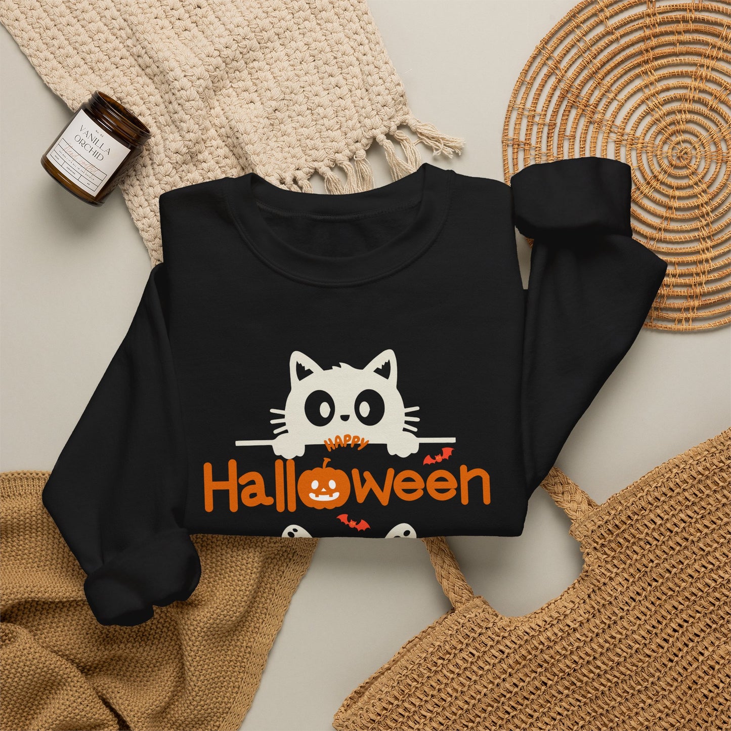 A black long-sleeve sweatshirt laid flat on a light beige surface, styled with a cream knit blanket, a small amber candle jar, and woven circular mats. The sweatshirt has a white cartoon cat peeking over orange text that reads 'HAPPY Halloween' with small orange bats and a pumpkin graphic.