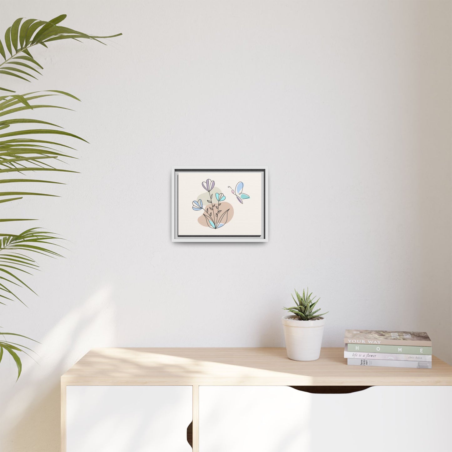 Framed artwork on a wall above a desk with a plant and books.