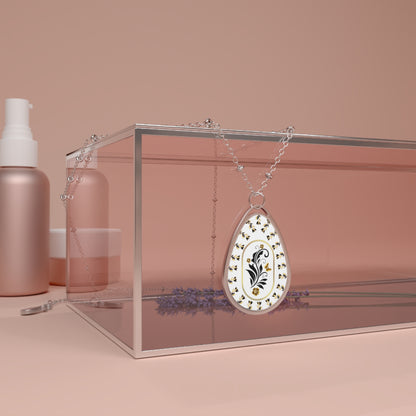 Teardrop-shaped pendant with floral design on a chain, placed inside a clear box with a pink background.