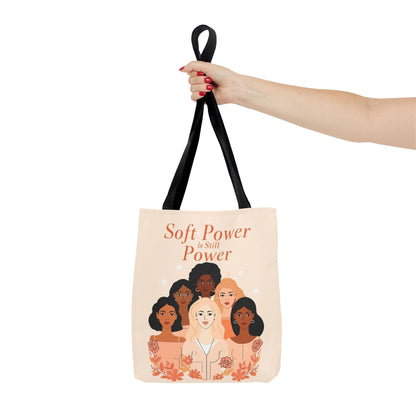 Tote bag with illustration of diverse women and text 'Soft Power is Still Power' held by a hand.