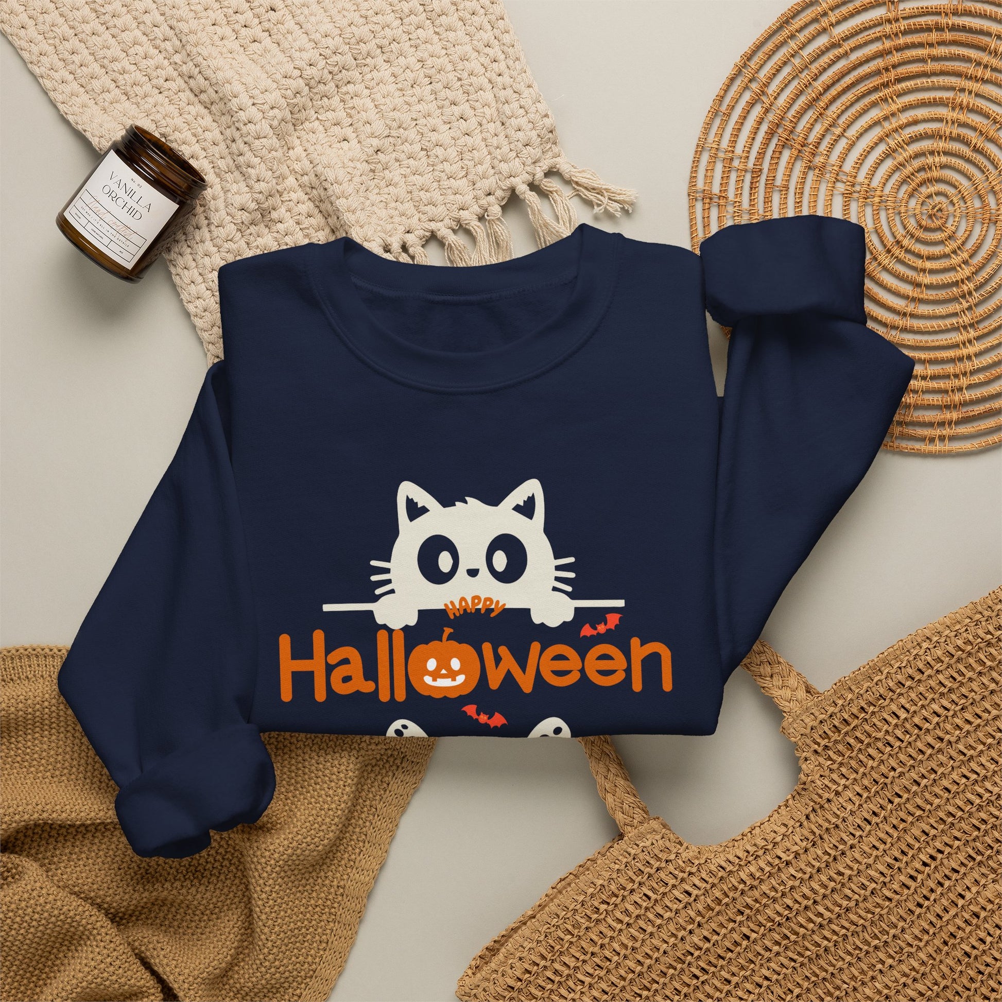 A navy blue sweatshirt with a cute white cat peeking over the text 'HAPPY Halloween' in orange, styled in a flat lay with a cream knit blanket, a woven mat, and a small candle.