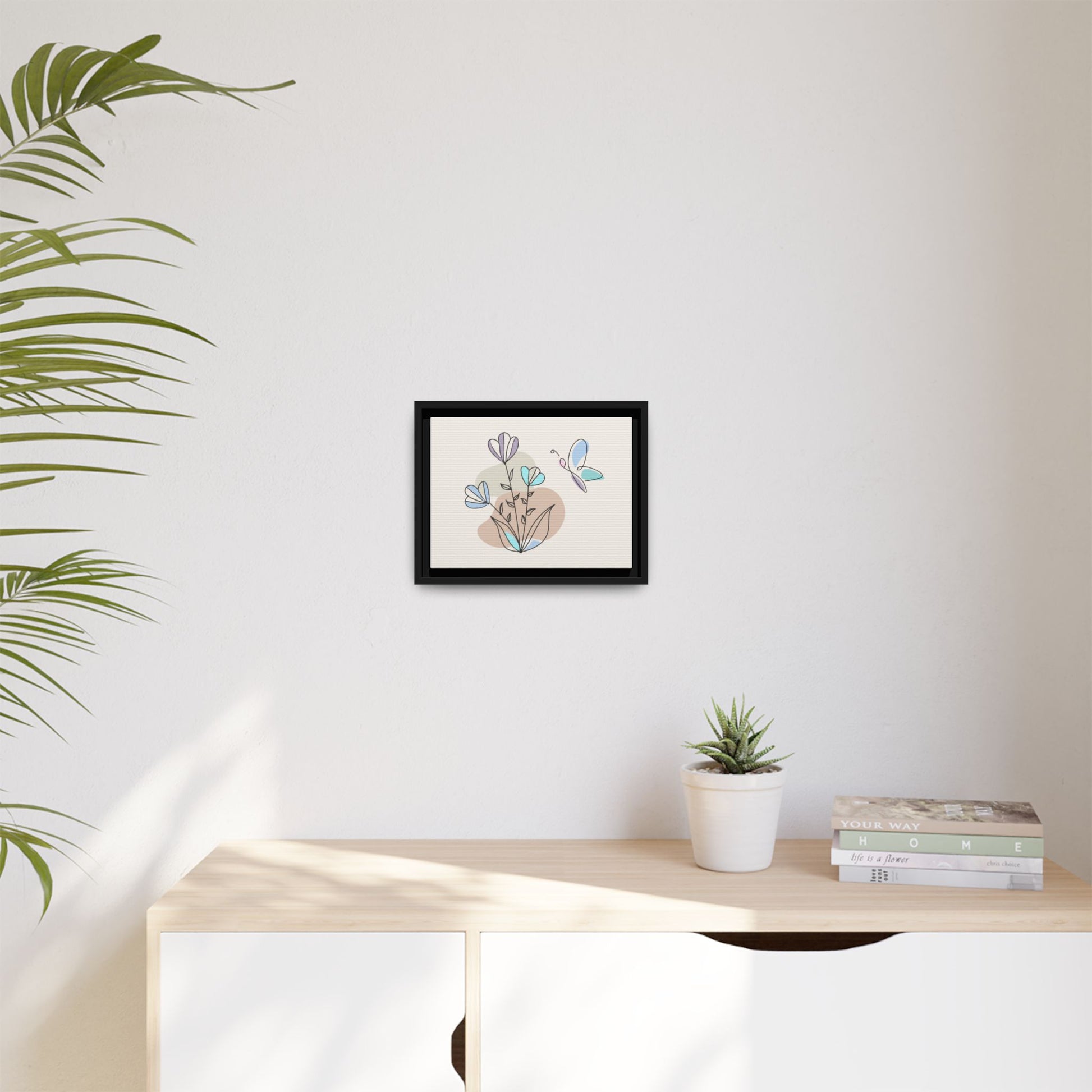 Framed artwork on a wall above a desk with a plant and books.