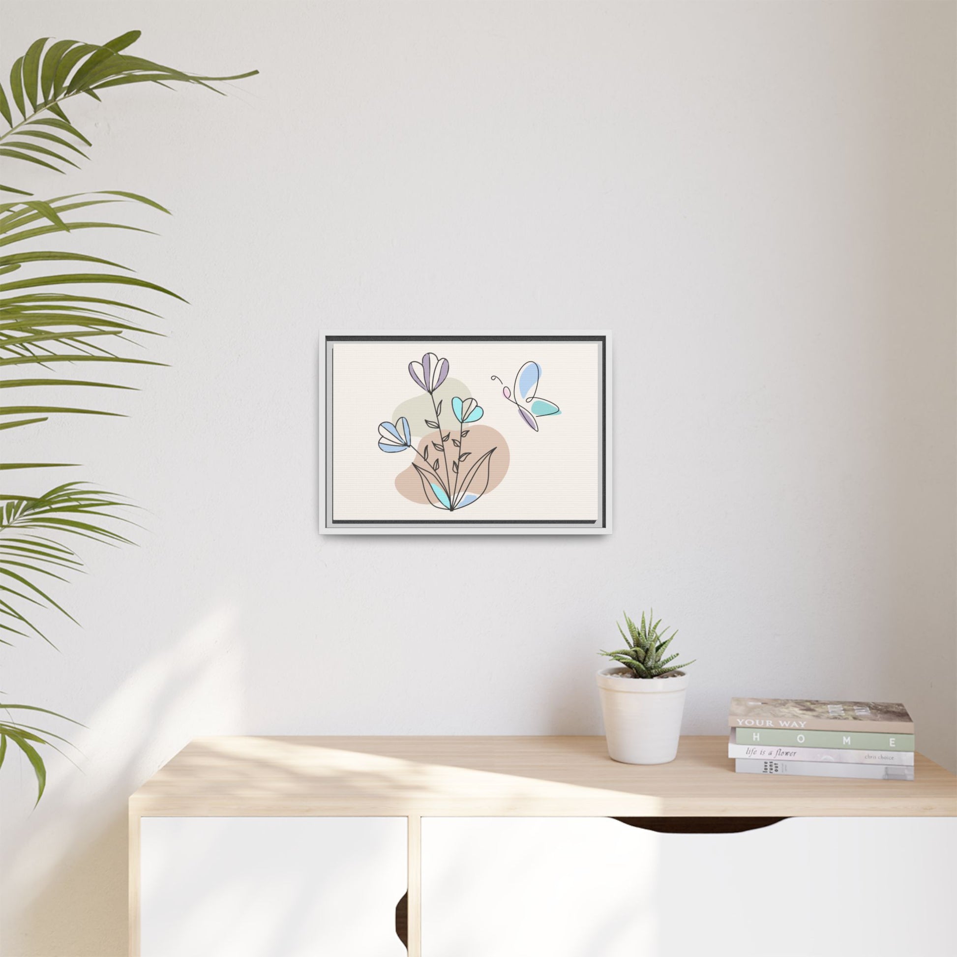 Framed artwork of flowers and a butterfly on a wall above a desk with a plant and books.
