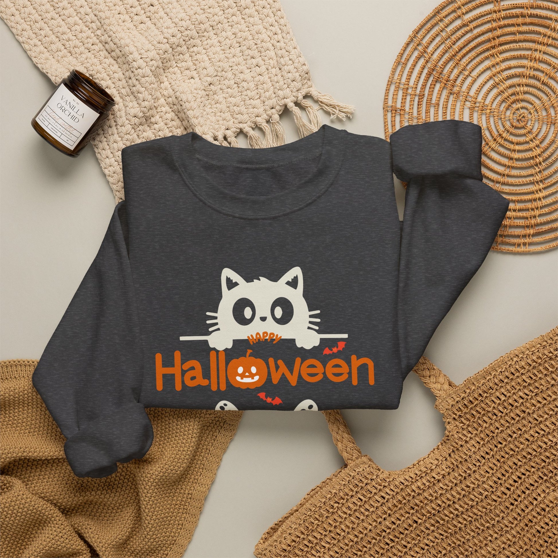 A dark charcoal gray sweatshirt with a 'HAPPY Halloween' graphic featuring a white cat and orange pumpkin details, styled in a flat lay with a woven blanket, a small candle, and a woven mat.