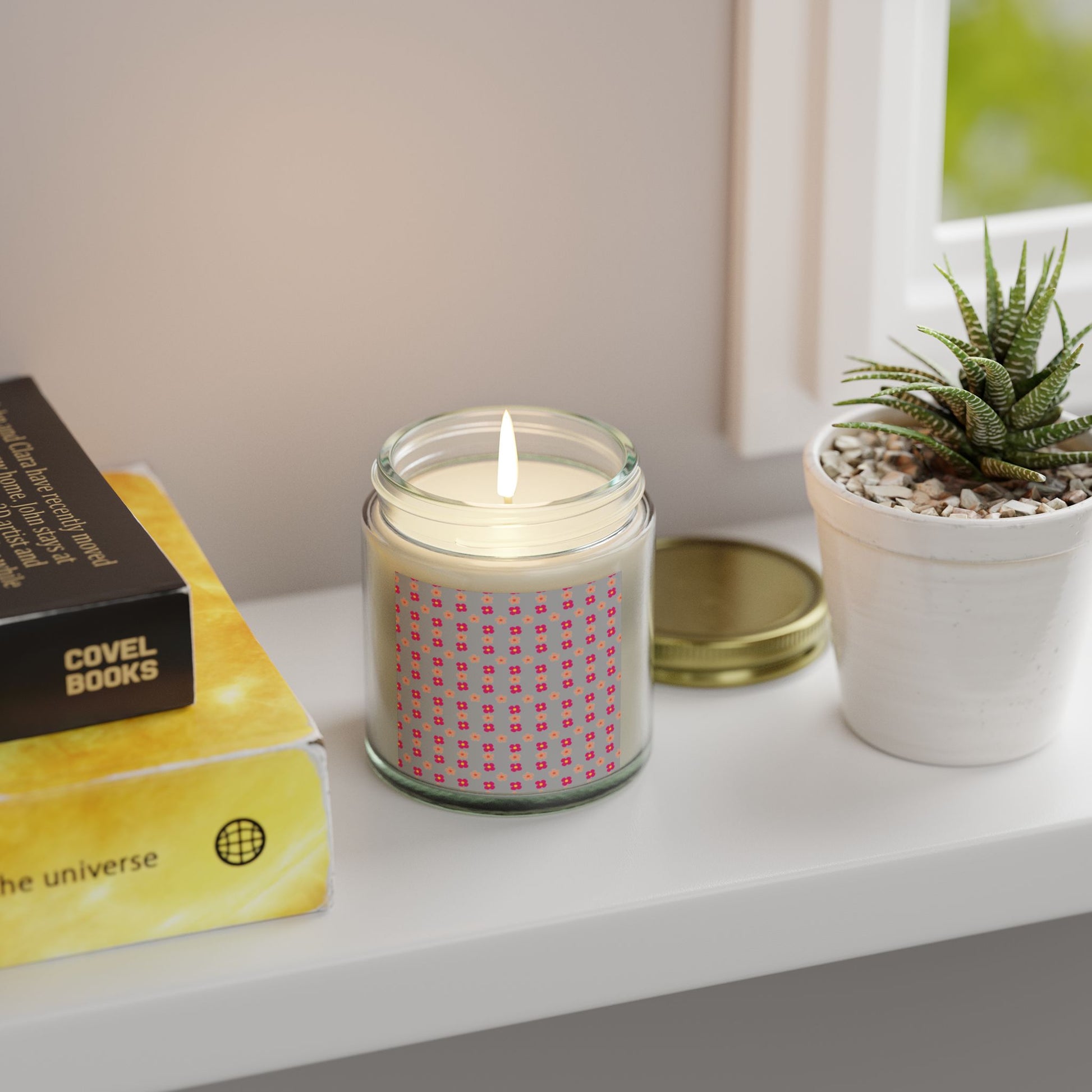 Candle in a glass jar on a shelf with a book and plant.

