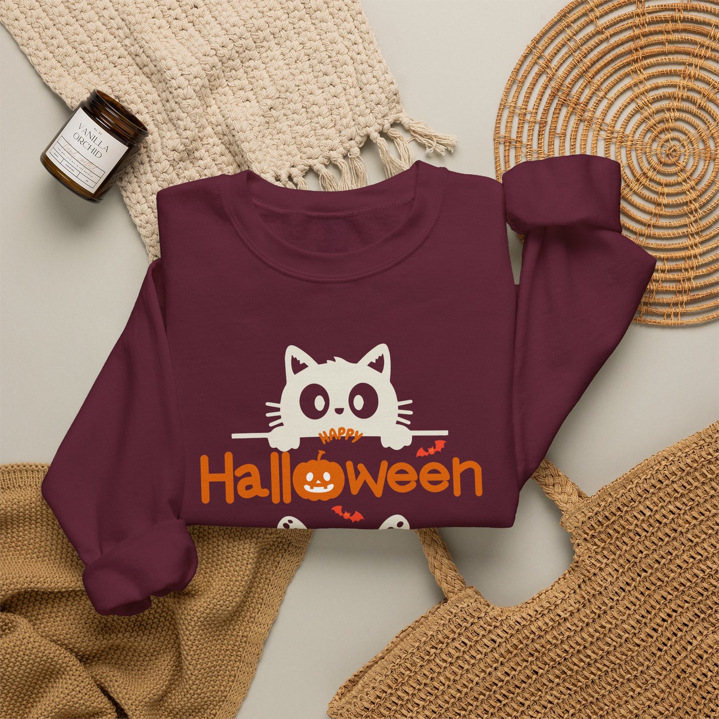 A folded maroon long-sleeve sweatshirt with a white cat peeking over orange 'Halloween' text, styled on a neutral surface with woven textiles and a small candle.