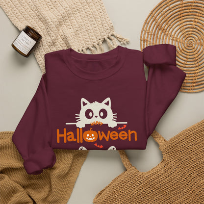 A folded maroon long-sleeve sweatshirt with a white cat peeking over orange 'Halloween' text, styled on a neutral surface with woven textiles and a small candle.