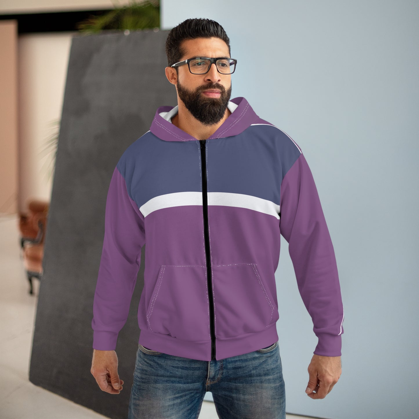A man wearing a purple and navy blue colorblock zip-up hoodie with a white chest stripe, paired with blue jeans, standing in an indoor setting.