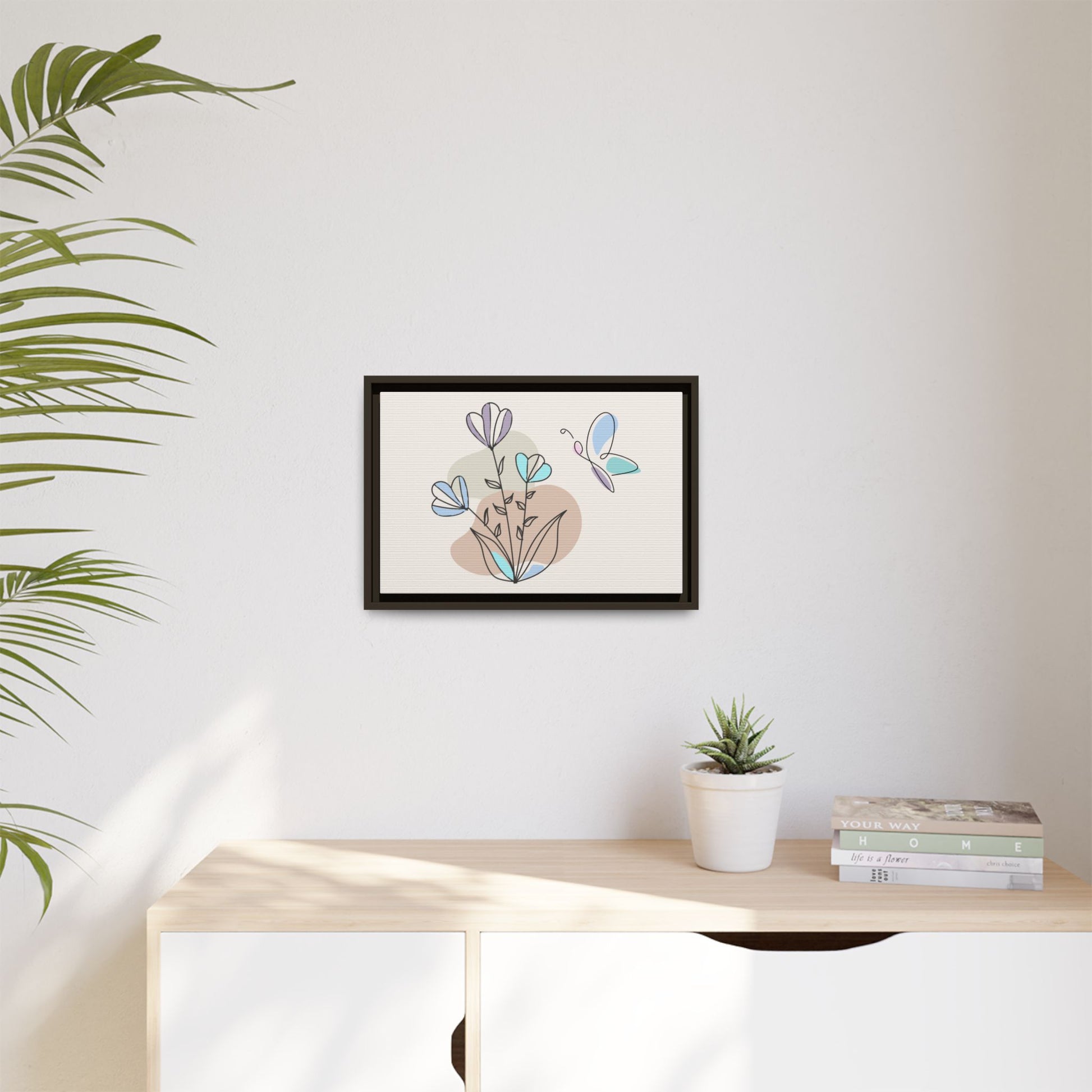 Framed artwork of flowers and a butterfly on a wall above a desk with a plant and books.