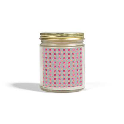 A scented candle with a gold lid and a label featuring a pink geometric pattern, placed against a plain background.