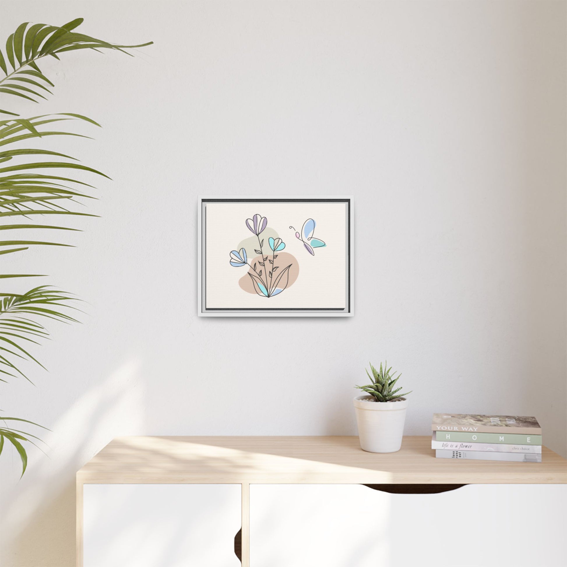 Framed artwork with flowers and a butterfly on a wall above a desk with a plant and books.