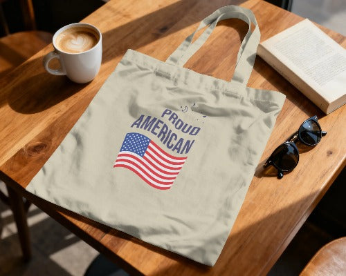 Tote bag with 'Proud American' text and American flag on a wooden table with a cup of coffee, book, and sunglasses.