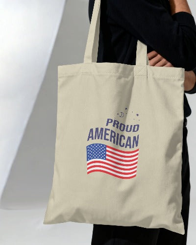 Beige tote bag with American flag and 'Proud American' text held by a person.